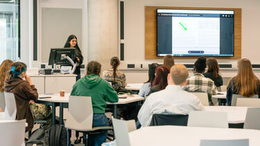 Student presenting at front of class, with other students sat at tables watching. 