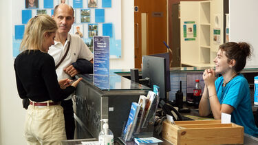 A female student and her father stood at the customer service desk talking to a smiley female member of staff