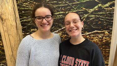 Two young women smiling at the camera standing in front of a nature poster