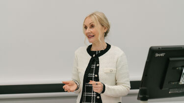 A woman with blonde hair and a white cardigan speaking, arms outstretched.