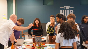 A group of students stood around a table with a large map on it, a man in a white chefs jacket holds a chilli pepper in his hand as he speaks to them