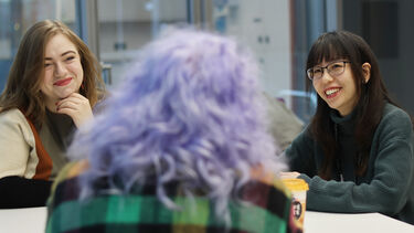 Three students sat around a table talking and smiling