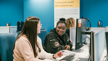 two students chatting in the Support Through Court room. In front of a PC in a cubical desk