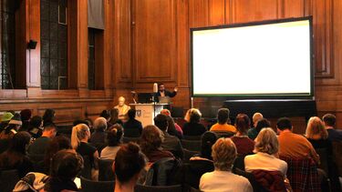 Gee at the front of the room with a PowerPoint slide on a projector. The audience is sat in front of the projector. 