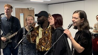 Five students singing behind 3 microphones. One plays a guitar