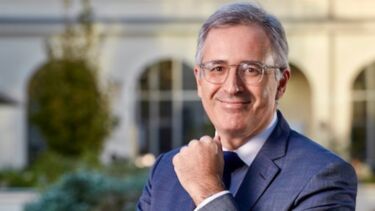 A man with grey hair, glasses and a navy suit smiling. 