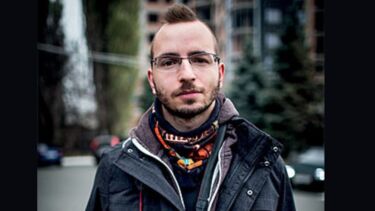 A man with black glasses, a leather jacket and a mohawk hairdo holding a camera.
