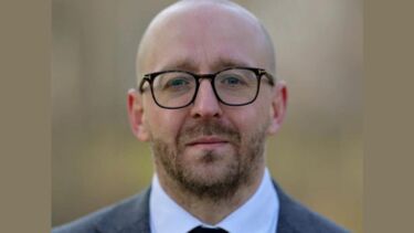 A bald man with stubble, wearing brown glasses and a blue suit jacket with a white shirt and tie. 