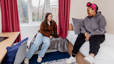 Two girls sat in a bedroom talking, one is on the bed and the other is on a window seat