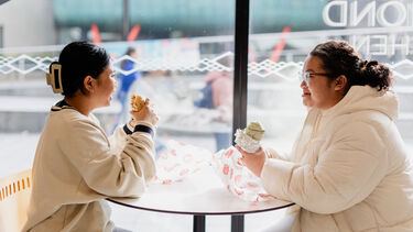 Two girls sat at a table in front of a large window eating burritos 