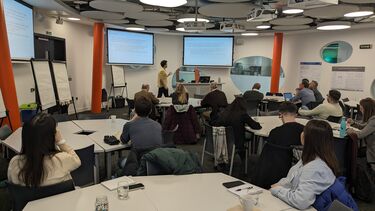 People sat at tables, watching a speaker present slides
