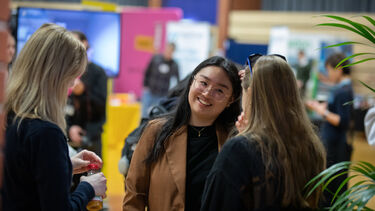 Three women having a discussion at an exhibition. The woman facing the camera is smartly dressed and smiling. In the background there are a number of other attendees and exhibition stands at the event.