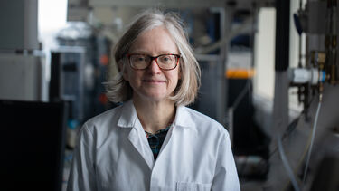 A portrait of Heather Walker, technician, in the foreground and wearing a lab coat, with a Biosciences lab in the background.