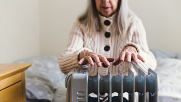 An older woman sits on a bed, holding her hands over a small electric heater.