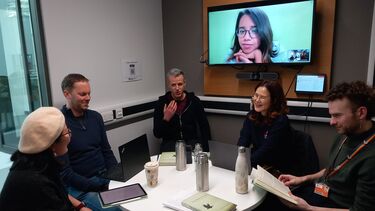Group of people sat around a table with books and laptops, in conversation 