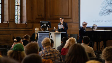 A speaker at a lectern, and a musician at a piano, in front of an audience. 
