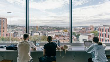 Three students sat at a desk with their backs to the camera. They're facing a view of the Sheffield metropolis from high up.