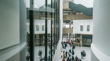 Interior of a large, bright building with several staircases and dozens of people walking in the foyer. 