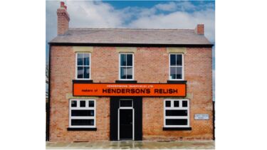 Red brick two storey house with chimney 3 upstairs windows and 2 downstairs windows either side of a black door.  A large rectangular orange Henderson's relish sign sits between the windows of the ground and first floors.