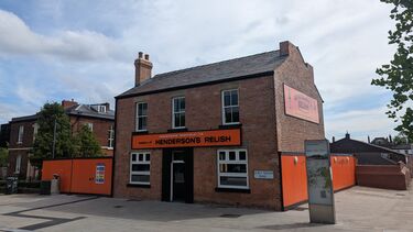 Photo taken from the corrner of a red brick two storey house with chimney, 3 upstairs windows and 2 downstairs windows either side of a black door.  A large rectangular orange Henderson's relish sign sits between the windows of the ground and first floors. Another orange Henderson's Relish sign is positioned on the end of the house at the same height at the first floor windows