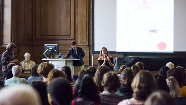 The backs of heads sat watching a person with a microphone 