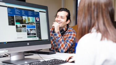 A man with dark, long hair and a orange checked shirt sitting in front of a computer screen. 
