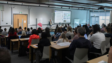 A man wearing a cream top and dark blue jacket stands at a lecture addressing a lecture theatre full of people.