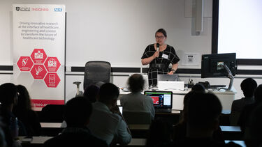A woman with dark hair tied back and dark rimmed glasses and wearing a black dress with a white grid pattern is stood at a lecturn holding a microphone addressing a lecture theatre audience.
