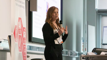 A woman with long, strawberry blonde hai waring a black cardigan and pale top r is stood speaking at a lectern in front of a presentation holding a microphone and gesturing with her other hand