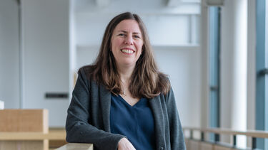 Claudia leans against a wooden balcony rail and is smiling at the camera 