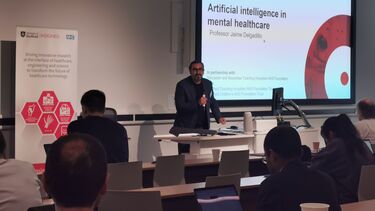A man with dark hair, dark glasses and a beard wearing a dark grey jacket and dark top is stood at a lectern holding a microphone in front of a presentation, looking at a lecture theatre audience.