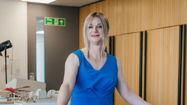 Jennifer leans on a glass balcony with her arms resting on the handle and smiles at the camera. 