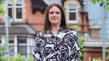 Julie stands in the wellbeing garden and is smiling at the camera with her hands folded in front of her.