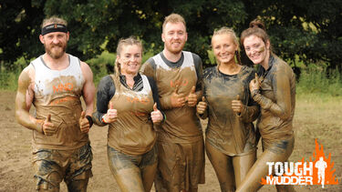 A group of people very muddy smiling to the camera with their thumbs up