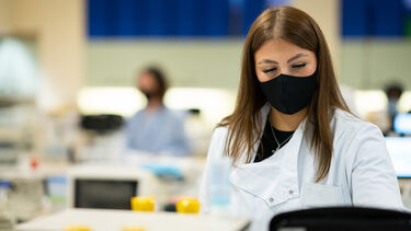 A woman in a lab coat and face mask works with laboratory equipment