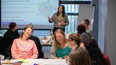 A presenter in the background with a classroom full of people around tables in the foreground