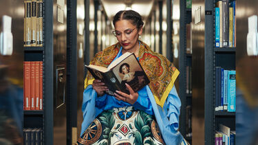 Terezia stands reading a book with a scarf around her shoulders 