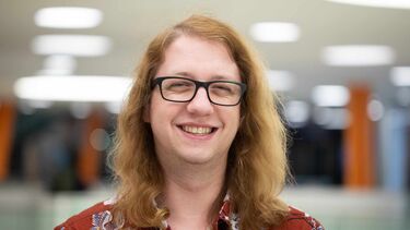An image of Jamie Howe, he looks directly into the camera and smiling. He is wearing a red shirt, glasses and has long ginger hair.