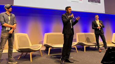 3 men presenting on stage with powerpoint screen on in background