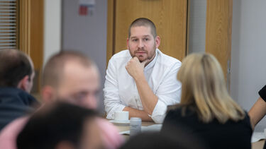A man in a white shirt sits at a table listening to discussions