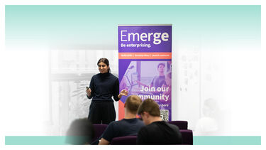 A woman presenting to a group in front of a banner that reads Emerge - Be enterprising