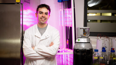 A person stands in a lab setting, in front of fluorescent lights, wearing a lab coat.