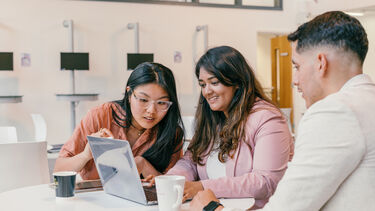 Three students sat at a table looking at a shared laptop screen