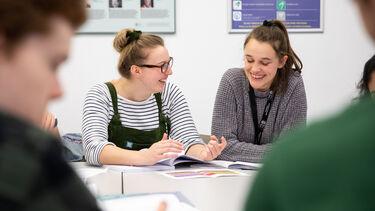 Two male students are in the foreground, blurred. Two women students are in focus; they are excitedly talking to each other. The students are taking part in a seminar in the Faculty of Health, University of Sheffield. The students are sitting around a grey table. 