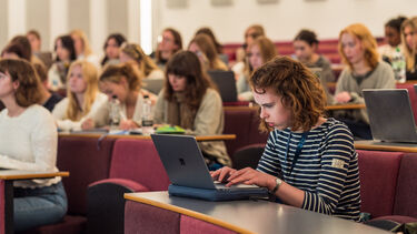 A group of students listening to a lecture, one is typing on their laptop.