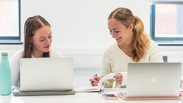 Two students in a lecture with laptops in front of them, they are smiling at eachother