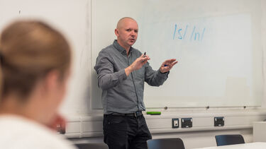 A lecturer stood at the front of a lecture theatre gesturing to the whiteboard