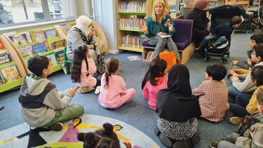 Children sat on the carpet in a library, listening to somebody read 