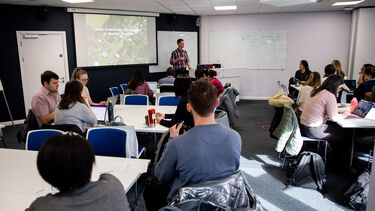 Students sitting around tables in a seminar room while a lecturer gives a presentation