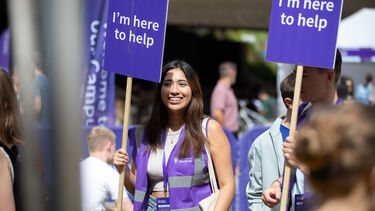 A smiling Sheffield student holding a sign that says "I'm here to help" at an undergraduate open day